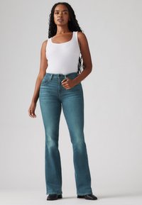 Calças de ganga de cintura alta e boca larga em denim verde-teal desgastado, com desgaste subtil, combinadas com uma blusa de alças branca com padrão rillet. Textura suave e corte ajustado.