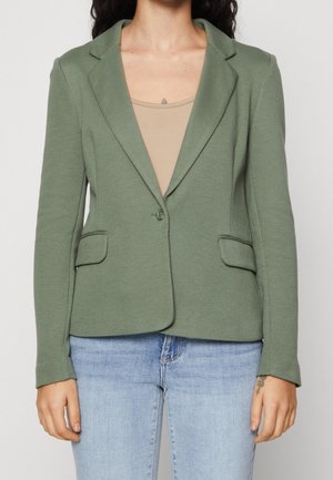 Kvinde iført en enkelt-knaps olivengrøn blazer over en beige top og lyseblå jeans, stående mod en ensfarvet baggrund.