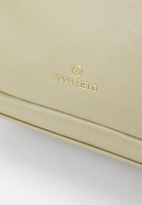 Gros plan d’un sac en cuir beige texturé avec le logo doré Anna Field et un emblème floral circulaire sur la surface.