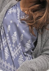 Modrý top s bílými geometrickými vzory, s kulatým výstřihem, spárovaný s dlouhým světle šedým kardiganem vyrobeným z jemného, texturovaného materiálu.