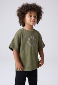 Olivgrön bomullst-shirt med korta ärmar, med ett ljusfärgat grafiskt tryck i ett cirkulärt mönster på framsidan. Svarta byxor.