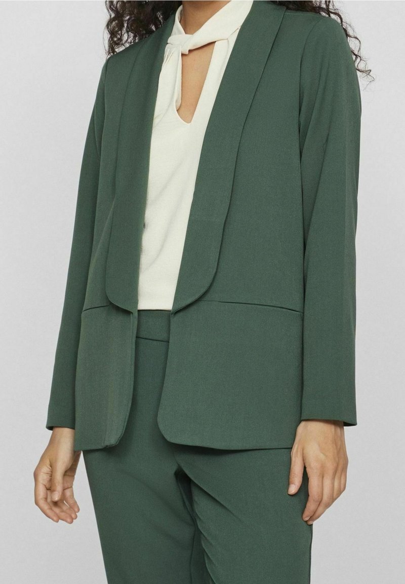 Femme portant un blazer vert foncé et un pantalon assorti sur un haut blanc avec un décolleté en forme de goutte et un détail noué au cou.