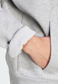Sweatshirt gris avec une manche côtelée blanche. La main se trouve dans une poche latérale, mettant en valeur la texture douce du tissu.