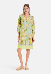 Blumendruck-Kleid in Pastellfarben mit Dreiviertelärmeln, V-Ausschnitt und strukturiertem Saum. Kombiniert mit beigen Rutschsandalen.