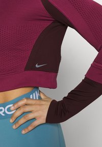 Top deportivo granate con paneles de malla, mangas largas y detalle de logo gris, combinado con leggings turquesa que llevan texto blanco en la cinturilla.