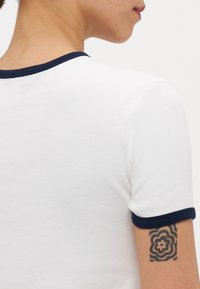 Hvit ribbet t-skjorte med marineblå ribbet kant på utkanten av halsen og ermene. Synlig tatovering på armen nær ermet.