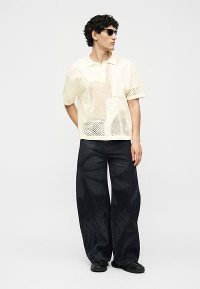 Άνδρας φορώντας γυαλιά ηλίου, κρεμ πλεκτό polo με κοντά μανίκια και σχέδια από δίχτυ, χαλαρό σκούρο παντελόνι με φαρδιά μπάσκα, στέκεται μπροστά σε μονόχρωμο φόντο.