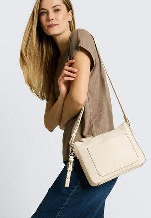 Frau mit langen blonden Haaren trägt ein taupefarbenes kurzärmeliges Oberteil und blaue Jeans, mit einer cremefarbenen Leder-Umhängetasche über der Schulter.