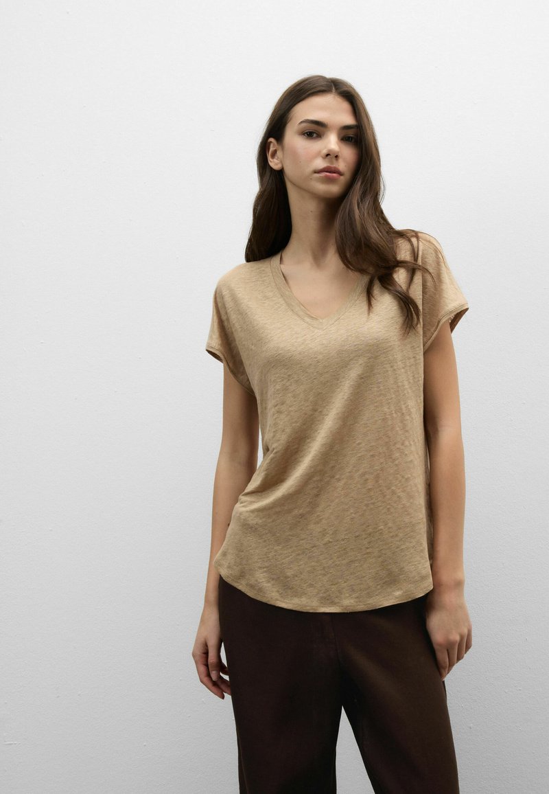 Femme aux longs cheveux bruns portant un haut beige à manches courtes et col en V, et un pantalon foncé, debout devant un mur gris clair uni.