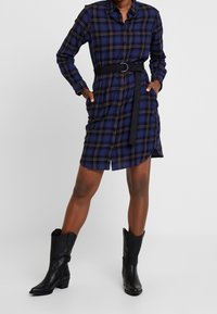 Marinblå rutmönstrad skjortklänning med bälte i midjan och långa ärmar, med knappar framtill och sidfickor, kombinerad med svarta lädercowboystövlar.