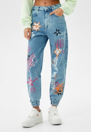 Pantalon jogger en denim avec des imprimés colorés d’étoiles, de fleurs et de textes, poignets élastiques, porté avec des baskets blanches à plateforme et un sweat court vert.