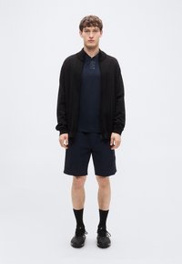 Μαύρο τζάκετ με φερμουάρ πάνω από μπλε σκούρο πόλο πουκάμισο, σε συνδυασμό με μπλε σκούρες βερμούδες. Το μοντέλο φοράει μαύρα αθλητικά παπούτσια και κάλτσες, στέκεται μπροστά σε απλό φόντο.
