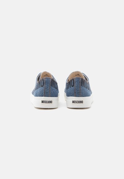 Ζευγάρι μπλε denim sneakers Moschino slip-on, εμφανιζόμενα από πίσω με λευκές σόλες και λογότυπο Moschino στην ταμπέλα της φτέρνας.