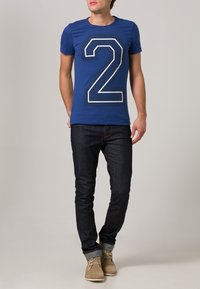 Blå kortärmad t-shirt med ett stort vitt nummer 2, matchad med mörka jeans och ljusbruna casualskor. Enkelt bakgrund.