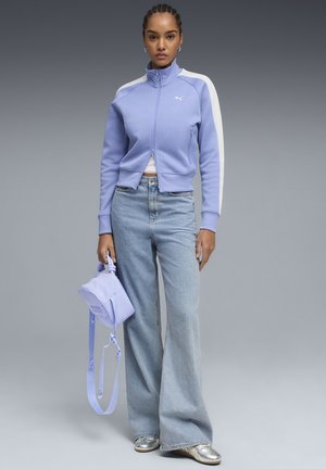 Mujer de pie con una chaqueta con cremallera de color púrpura claro con rayas blancas, vaqueros de pierna ancha azul claro, zapatos plateados y sosteniendo un bolso púrpura a juego.