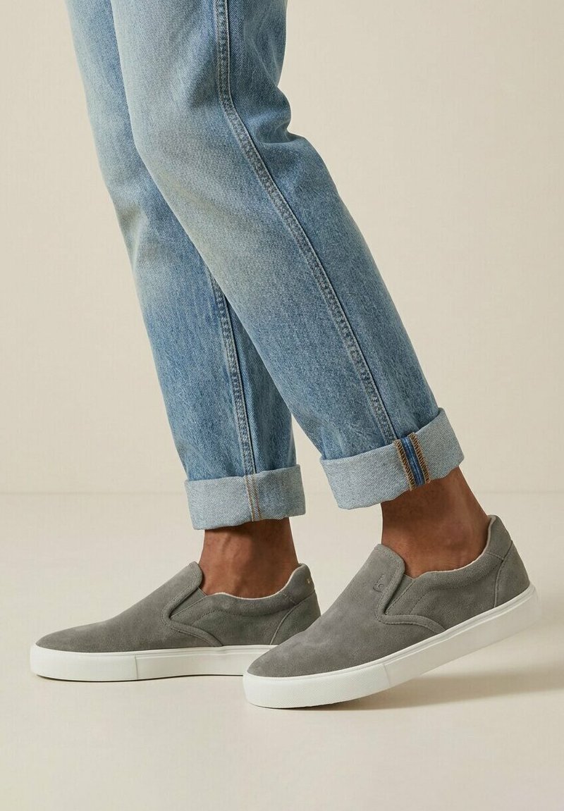 Fødder iført grå slip-on ruskindssneakers med hvide såler, kombineret med opsmøgede lyseblå cowboybukser mod en ensfarvet baggrund.