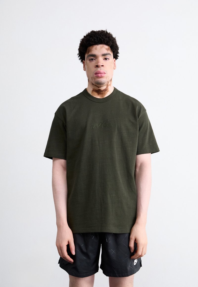 Oliven grøn bomulds T-shirt med korte ærmer, der har et subtilt broderet logo på brystet, parret med sorte shorts med et geometrisk mønster.