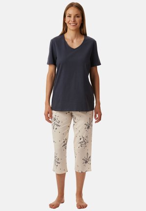 Mujer sonriente de pie descalza, con una camiseta de manga corta y escote en V de color oscuro y pantalones capri claros con un pequeño estampado floral oscuro.