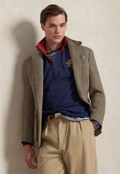 Καφέ σακάκι με σχέδιο houndstooth φορεμένο πάνω από μπλε σκούρο πόλο με κόκκινο γιακά. Μπεζ παντελόνι και ασημένιο ρολόι ολοκληρώνουν την εμφάνιση. Ορατές λεπτομέρειες υφής υφάσματος.