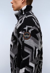 Giacca in pile con un motivo geometrico in bianco, grigio e nero. Collo alto e patch con logo sul braccio sinistro. Tasca laterale con zip.