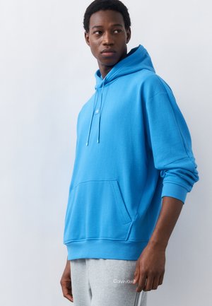 Jaunas vyras vilki ryškiai mėlynį džemperį su priekine kišene ir pilkus Calvin Klein laisvalaikio kelnes, stovintis priešais paprastą šviesią foną.