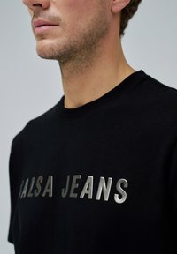 Camiseta negra con un logotipo metálico plateado de "SALSA JEANS". La toma en primer plano muestra la textura de la tela y los detalles del cuello.