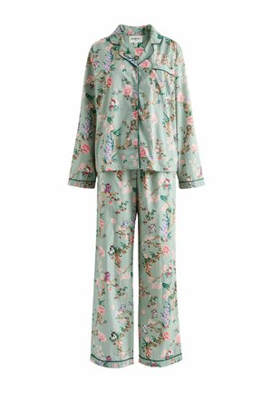 Kobieta stojąca w kwiecistym, jasnoniebieskim komplecie piżamy z długimi rękawami i spodniami, na tle różowo-fioletowej przezroczystej tkaniny.