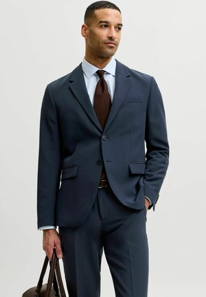 Homme en costume bleu marine et cravate marron tenant un sac en cuir marron, debout devant un fond gris clair uni, regardant légèrement sur le côté.