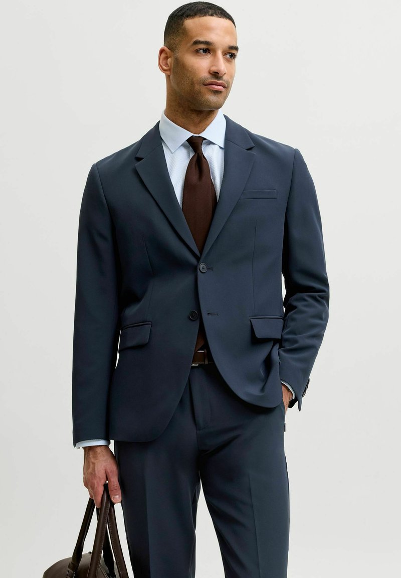 Homme en costume bleu marine et cravate marron tenant un sac en cuir marron, debout devant un fond gris clair uni, regardant légèrement sur le côté.