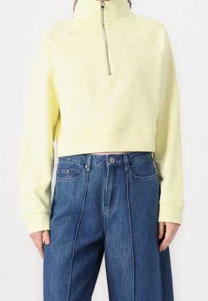 Kobieta nosząca bladożółtą krótka bluzę z zamkiem błyskawicznym z przodu oraz niebieskie dżinsy z wysokim stanem i pionowymi szwami.