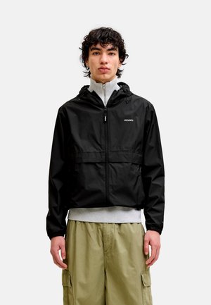 Mladić s kovrčavom crnom kosom, odjeven u crnu jaknu na patentni zatvarač s kapuljačom preko svijetlosivog džempera i bež cargo hlača, stoji ispred bijele pozadine.