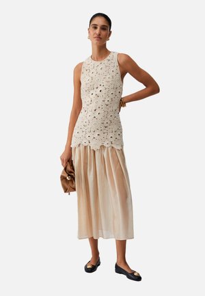 Donna che indossa un top beige senza maniche lavorato all'uncinetto, gonna midi beige chiaro, ballerine nere con dettaglio dorato, tiene una pochette marrone, in piedi con una mano sul fianco.