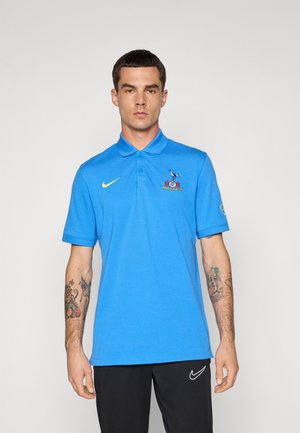 Mand med kort mørkt hår iført en blå Nike polo med et fugleemblem på brystet, sorte bukser med Nike-logo og har tatoveringer på begge underarme.