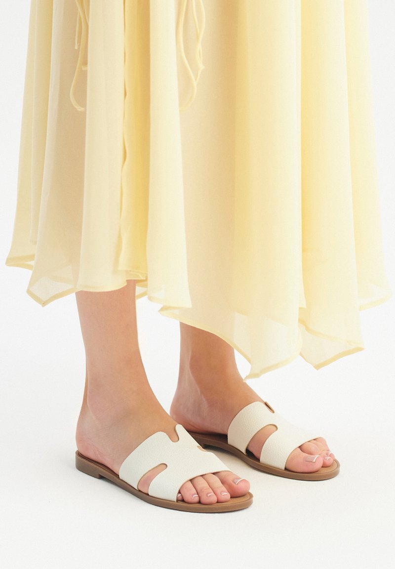 Sandali bianchi slip-on realizzati in materiale testurizzato, con disegni ritagliati, suola piatta e dettagli in marrone chiaro. Abbinati a un vestito giallo trasparente.