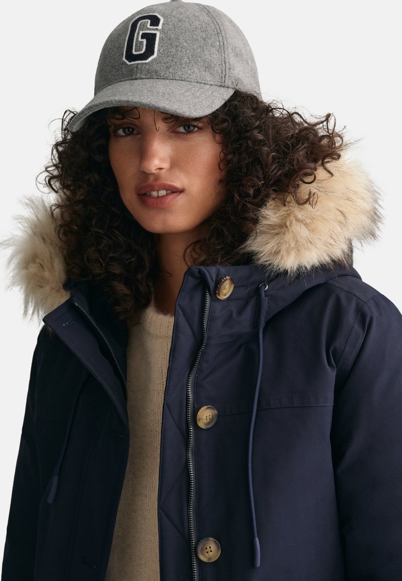 Marine Parka mit einer Kapuze aus Kunstfell, Knopfverschluss und einem hellbeigen Pullover darunter. Graue Mütze mit schwarzem Buchstaben "G".