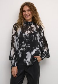 Chiffon blouse met zwart-wit bloemenpatroon, geplooide textuur, hoge hals met ruffles en lange mouwen. Gepaard met zwarte broeken.