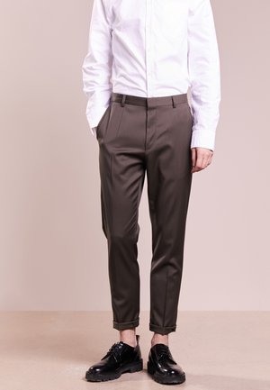 Hombre vestido con pantalones marrones a medida con puños, zapatos de vestir negros pulidos y una camisa blanca abotonada, de pie frente a un fondo liso.