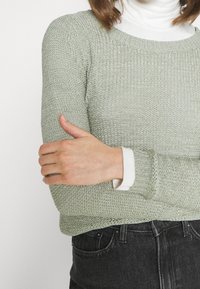 Jasnozielony sweter z dzianiny o teksturowanym wykończeniu, okrągłym dekolcie i ściągaczami na rękawach, nałożony na biały golf. Połączony z ciemnymi dżinsami.