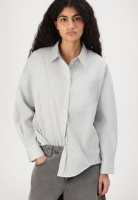 Camicia a maniche lunghe a righe in grigio chiaro e bianco, con chiusura a bottoni sul davanti, colletto e una tasca sul petto. Morbida, con texture intrecciata.