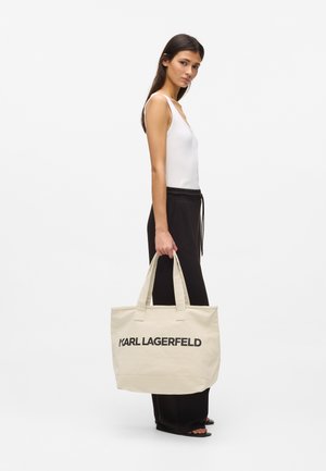 Jeune femme portant un débardeur blanc et un pantalon noir, tenant un sac cabas beige Karl Lagerfeld à son côté, sur un fond blanc uni.
