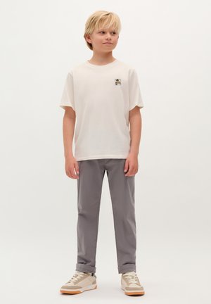 T-shirt bianca in cotone con maniche corte e un piccolo patch grafico, abbinata a pantaloni grigi chiari e sneakers color beige con un motivo testurizzato.