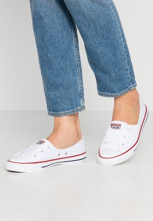 Person iført blå jeans og hvide lave Converse-sneakers står på et lyst gulv mod en ensfarvet baggrund.