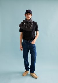 Schwarzes Kurzarmhemd mit grauem Schal, blaue Jeans und beigen Slipper; marineblauer Hut mit weißem Logo; steht vor einem hellblauen Hintergrund.