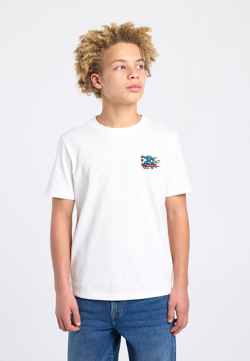 Tienerjongen met krullend haar, gekleed in een wit T-shirt met een kleine kleurrijke skateboardafbeelding en een blauwe spijkerbroek, tegen een effen achtergrond.