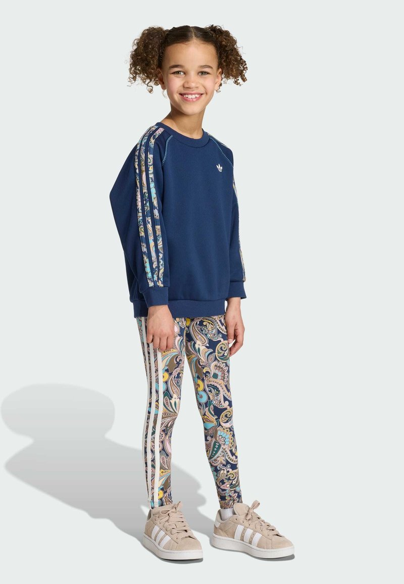 Jeune fille aux cheveux bouclés porte un sweat-shirt bleu marine avec des rayures à motifs et des leggings assortis à motif cachemire, souriante et debout sur un fond clair.