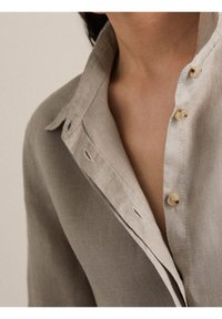 Chemise en lin gris clair avec col pointu, fermeture boutonnée sur le devant et détails de couture subtils. Texture lisse et design minimaliste.