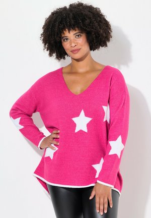 Rosa Strickpullover mit V-Ausschnitt und langen Ärmeln, mit weißen Sternmustern. Der Saum ist hinten länger, strukturierter Stoff. Kombiniert mit schwarzen Hosen.