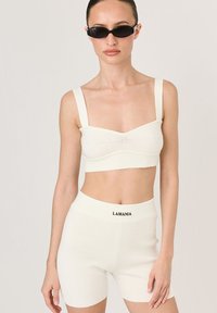Biały prążkowany crop top z szerokimi ramiączkami, w połączeniu z dopasowanymi szortami o wysokim stanie z nazwą marki "LAMANIA" w kolorze czarnym.
