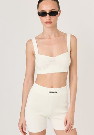 Biały prążkowany crop top z szerokimi ramiączkami, w połączeniu z dopasowanymi szortami o wysokim stanie z nazwą marki "LAMANIA" w kolorze czarnym.