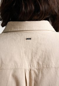 Lichtbeige overhemd van een licht gestructureerde stof, met een geplooid ontwerp aan de achterkant en een klein metalen logootje nabij de kraag.